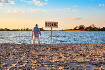 Warning sign with inscription closed along the sand beach of the river and male wearing protective suit stands with his back. Contaminated water, quarantine, virus outbreak, crime scene concept
