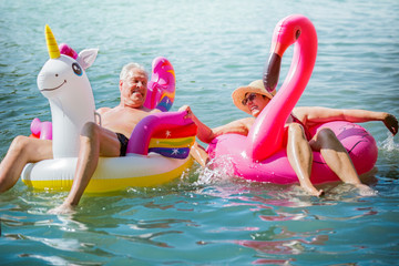 Elderly couple having fun on inflatable flamingo and unicorn. Funny active pensioners happy...