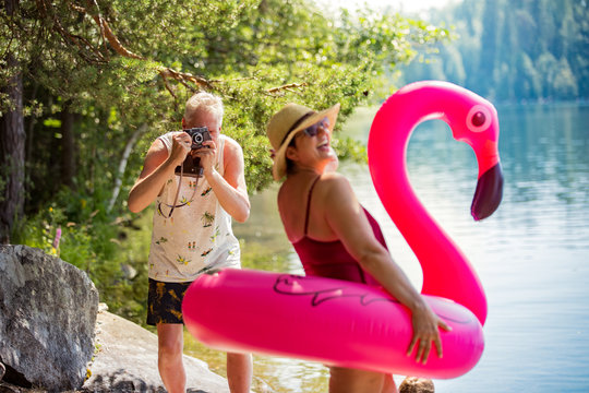 Elderly couple taking photos on vintage camera in forest along the seashore holding giant inflatable flamingo. Funny active pensioners enjoying summer vacation on the beach
