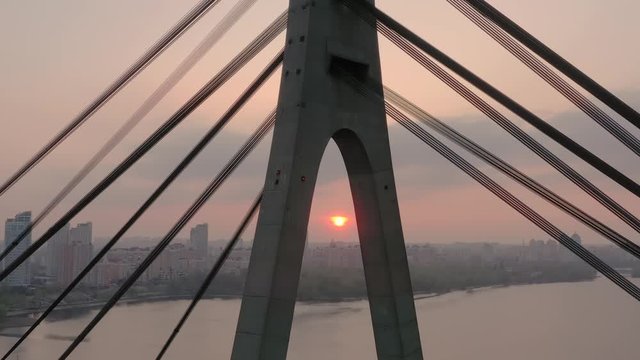 Aerial drone view View Of North or Moscow Bridge Kiev, Ukraine over Dnieper. Drone span between the cables of the bridge. Sunset sun. City view. the sun between the pylons of the bridge.