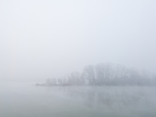 Peaceful autumn scene of mystical river island with a deciduous grove in deep fog early in morning. Calm and silent nature scene covered with thick fog.