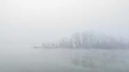 Peaceful autumn scene of mystical river island with a deciduous grove in deep fog early in morning. Calm and silent nature scene covered with thick fog.