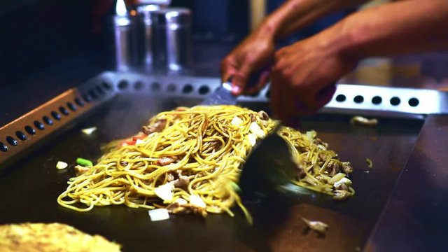 Chef mixes noodles, vegetable and flambeed chicken strips for Modan-yaki food (Okonomiyaki with yakisoba noodles) or Chicken and Vegetables Stir Fry spaghetti on grill in professional Asian kitchen.