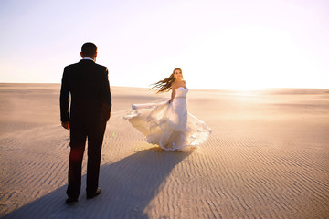 Fototapeta na wymiar The bride and groom are walking in the desert, blue sky and white sand, a beautiful couple, wedding day, the bride having fun and spinning on the dunes, a beautiful evening and sunset in the wild