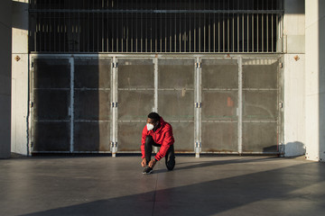 Young man on urban running workout lacing shoes. Black athlete training in the city with face mask against Covid-19.