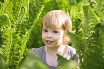 little emotional girl smiling playing in beautiful green thickets of fern, hiding behind large leaves. girl playing hide and seek in beautiful thickets. happy childhood