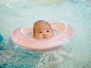 A cute 3 months little Asian baby girl learning to swim with swimming ring in an indoor pool, with copy space.