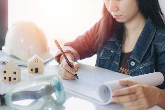 Asian Woman Checking Architectural Plan With Personal Safety Equipment And Wooden House Model On White Office Desk For Building Inspection Concept