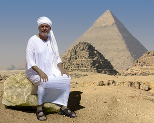 An ancient Egyptian sits on a rock in front of the historic Giza pyramids in Egypt. Right behind him are two little crumbled pyramids. The man is wearing a white robe and a turban.