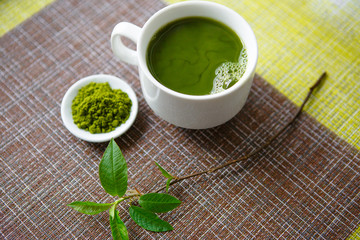 Japanese matcha green tea is poured into a white mug and on a white saucer in powder. Tea set on a textured napkin of natural flowers, decorated with a branch of green leaves. Background with space fo