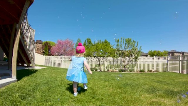 Litte girl toddler in a princess dress chasing soap bubbles in the backyard - slow motion