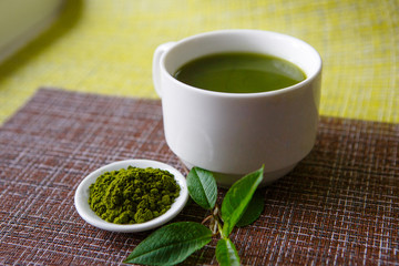 Japanese matcha green tea is poured into a white mug and on a white saucer in powder. Tea set on a textured napkin of natural flowers, decorated with a branch of green leaves. Background with space fo