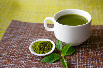 Japanese matcha green tea is poured into a white mug and on a white saucer in powder. Tea set on a textured napkin of natural flowers, decorated with a branch of green leaves. Background with space fo
