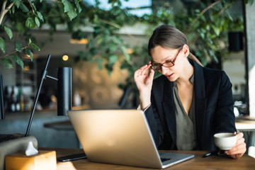 Business Woman with cup of coffee and laptop at table in a cafe. Focused business woman in stylish glasses sit on cafe working on laptop.