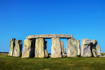 Closeup of Stonehenge standing stones with three lintels across four huge uprights in the foreground under a very blue sky on a summer day with a tilled field in the background