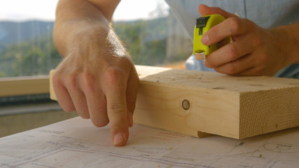 CLOSE UP, DOF: Builder points at his plans before measuring a block of wood.