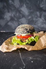 It is a black burger with meat and vegetables on the dark background. Fast food.