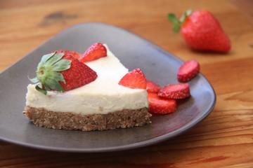A slice of cheesecake with strawberries on dark triangle plate and wooden table. Closeup, macro, horizontal.
