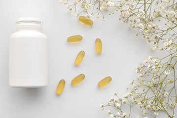 Obraz na płótnie Canvas Oil yellow gelatin capsules with white bottle on white background with flowers, vitamins and antioxidant concept Omega 3, liver cod or evening primose oil for healthcare. Minimalism. Copy space.