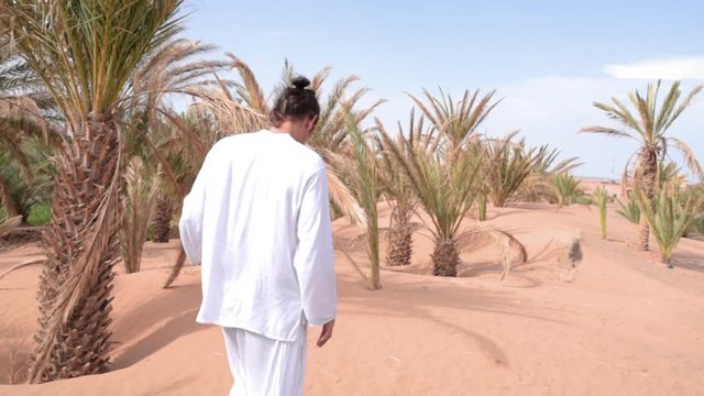 Stylish young man wearing white clothes walks on sandy Moroccan desert across beautiful palm trees 