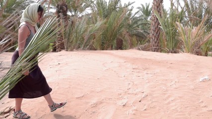Young woman barely walking through sandy dunes on desert of Morocco surrounded by palm trees on a hot day