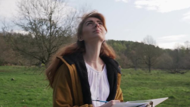 Young woman with notes in hand walks on path on nature trail and looks at the sky to see if there is something to discover