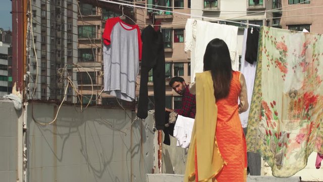 Indian woman hangs clothes on clothesline in suburbs and man stares at her, roof