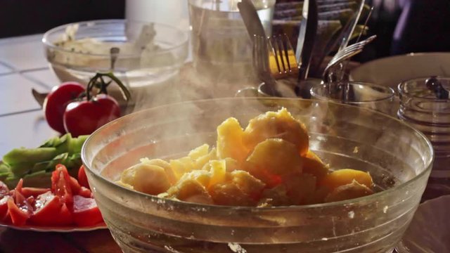 Static view of freshly boiled potatoes steaming in a bowl in a kitchen with fresh tomatoes and salad in the background