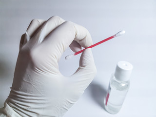 A hand in a white rubber glove holds a cotton swab against the background of a small bottle with an antiseptic. Image with selective focus