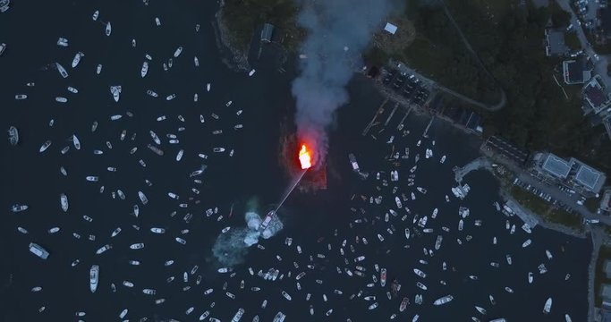 Extremely unique, high altitude, top down, turning drone footage of small motor boats and sail boats gathering around and witnessing the biggest bonfire event on Earth at Alesund, Norway at dusk.