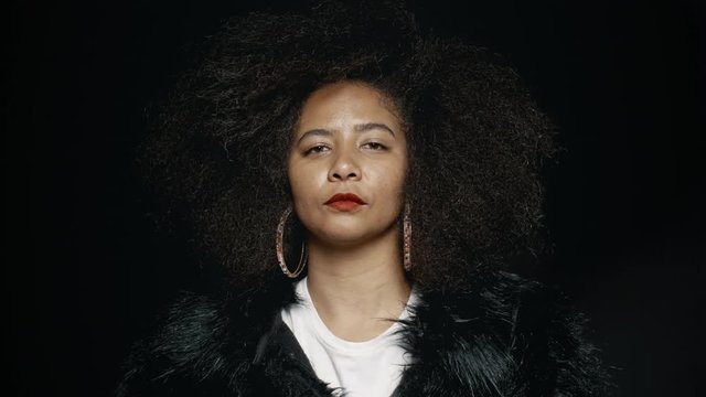 Mature woman with afro hairstyle stroking her hair against black background. African female in fur coat caressing her hair.
