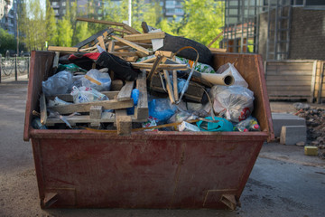 a litterless metal container without a cover is full of construction debris and waste standing in the middle of the street next to a building under construction in the city in the sun
