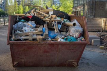 a litterless metal container without a cover is full of construction debris and waste standing in the middle of the street next to a building under construction in the city in the sun