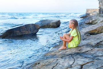 A little girl is sitting on the rocks by the sea. Long braided hair. Seashore. Gaze into the distance. Great stones. Summer evening. Dream good dreams. It's good to be alone.