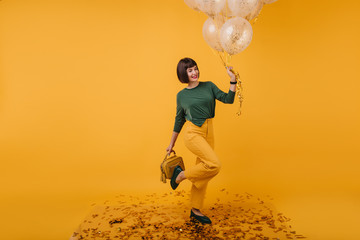 Fototapeta na wymiar Indoor portrait of birthday girl posing on one leg and smiling. Cute white woman in yellow pants dancing in studio.