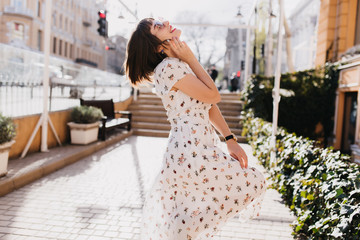 Naklejka premium Outdoor shot of graceful girl relaxing in sunny day. Stylish short-haired lady in white dress dancing on the street.