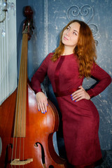 The girl in the red dress standing in the Studio holding a musical instrument the double bass.