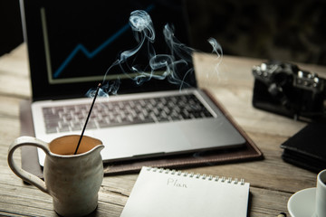  incense and blured laptop, notepad, growth chart, coffee, pen, leather wallet on a old gray wooden background. concept: businessman workplace