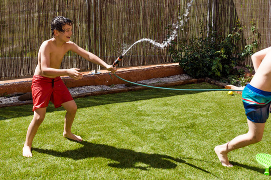 Two Boys Having A Water Battle On A Hot Sunny Day In  The Back Yard With A Hose.