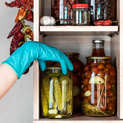 Homemade vegetables in jars on wooden shelves in the home pantry. Marinate food, as stocks from the autumn harvest, will be useful for self-isolation. Hand in blue glove get one can with pickles.