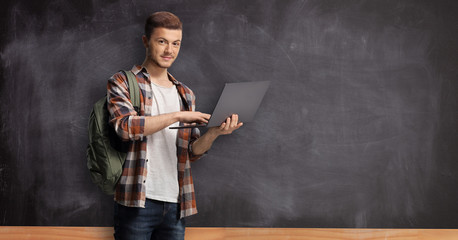 Male student working on a laptop and standing in front of a school blackboard