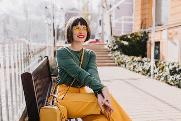 Refined young woman with short hair sitting on bench and smiling. Outdoor photo of amazing caucasian girl enjoying good spring day.