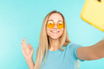 Young woman makes selfie over isolated blue background. Happy young lady makes OK sign and smiles.
