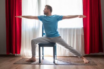 Caucasian young man in Warrior 2 Pose using chair at his living room