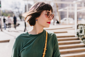 Portrait of pretty white girl in earrings looking around during street photoshoot. Outdoor photo of ecstatic short-haired brunette woman in sunglasses.