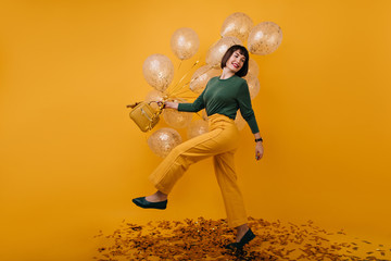 Fototapeta na wymiar Full-length portrait of adorable woman dancing in studio with party balloons. Indoor shot of blissful brunette girl in yellow pants having fun in her birthday.