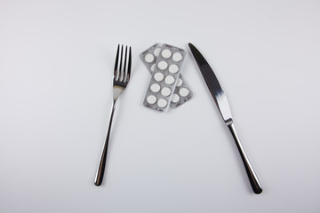 Iron fork and knife with pills on a white background. Use of medications. Care of health. Excessive use of medications during treatment. Eating pills.