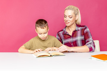 Attractive young woman and her little son are sitting at the table and doing homework together. Mother helps son with his school classes.