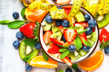 Concept of healthy lifestyle, dieting, slimming for summer. Colorful fresh delicious fruit salad in a bowl and tape measure. Wooden background, top view, flat lay, copy space for text