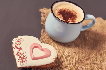 Blue mug of coffee with foam on sackcloth near gingerbread cookie in shape of heart on concrete surface. Concept of love or christmas. Toned and vintage image